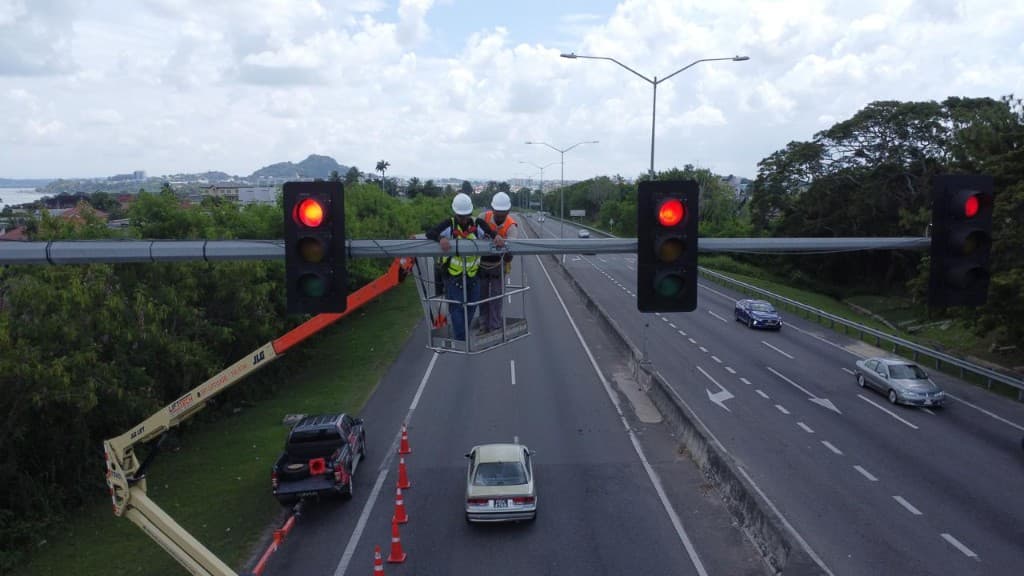 Technicians in a boom lift servicing overhead traffic signals above a highway with cones marking the work zone
